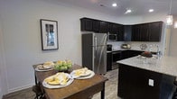 The Tracery offers a thoughtfully designed single-story layout that blends functionality with style. The kitchen serves as a central highlight, featuring rich, dark cabinetry that extends to the upper walls, creating a striking contrast against the light-toned walls and wood-look flooring. Stainless steel appliances, including a full-size refrigerator, microwave, and range, lend a modern, polished aesthetic to the space. Granite countertops add an upscale touch, complementing the kitchen island that provides additional prep and storage space. Recessed lighting throughout the kitchen ensures the area remains bright and well-lit. Adjacent to the kitchen, a dedicated dining area accommodates a substantial wooden dining table, ideal for both casual meals and entertaining. Crown molding along the ceiling adds an elegant architectural detail that elevates the overall ambiance. The wood-look flooring flows seamlessly between spaces, contributing to a cohesive and clean aesthetic throughout. The open connection between the kitchen and dining areas enhances the sense of space, making this home feel both welcoming and practical.