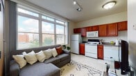 This 2-bedroom, 1-bathroom E Style unit is a well-organized single-floor residence designed for comfortable urban living. The open-concept living and kitchen area serves as the heart of the home, where a large multi-pane window floods the space with natural light and frames sweeping city views. Track lighting overhead adds a modern touch, while light-toned tile flooring ties the living and kitchen zones together seamlessly. The kitchen is equipped with rich, warm-toned wood cabinetry extending both above and below the counters, offering generous storage. White appliances — including a refrigerator, electric range, over-the-range microwave, and dishwasher — provide all the essentials for everyday cooking. Both bedrooms are carpeted and comfortably sized, each featuring windows that invite natural light and open onto urban streetscape views. The rooms offer a calm, neutral palette ideal for rest and personalization. The full bathroom features clean tile flooring, a glass-enclosed shower stall, and a simple, functional layout with ample towel storage. The overall ambiance of this unit is practical and welcoming, making it a solid choice for those seeking a functional urban residence with connected living spaces and city-facing outlooks throughout.