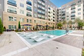 Located at 2120 Dunavant St in Charlotte's South End neighborhood, this outdoor amenity space is a meticulously designed courtyard retreat set within a modern multi-story building complex. The centerpiece is a generously sized swimming pool with crystal-clear blue water, surrounded by light-toned stone and wood-look deck pavers that lend a polished, upscale aesthetic. A distinctive sun shelf runs along one edge of the pool, outfitted with contemporary white lounge chairs that sit partially submerged — ideal for relaxing in the water. The pool deck is lined with additional chaise lounges and navy blue market umbrellas, providing ample options for sunbathing and outdoor leisure. A striking stone water feature wall anchors one end of the space, adding a tranquil visual and auditory element to the courtyard ambiance. Manicured Italian cypress trees and lush plantings are thoughtfully positioned throughout, offering greenery and structure to the design. Shaded pergola seating areas provide comfortable retreats from the sun, while a dedicated outdoor grilling station with a full-size gas grill and adjacent dining furniture makes alfresco entertaining effortless. The entire space is framed by the building's multi-story brick and panel façade, creating an intimate and resort-like atmosphere in the heart of Charlotte.