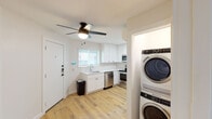 This single-floor unit has been thoughtfully renovated with a clean, modern aesthetic carried consistently throughout every room. Light hardwood-style flooring runs across the entire space, lending warmth and visual continuity from entry to bedroom.

The kitchen is a standout feature, outfitted with crisp white shaker cabinetry extending to the ceiling for ample storage, bright white countertops, and a full suite of stainless steel appliances including a refrigerator, electric range, over-the-range microwave, and dishwasher. A single-basin sink sits beneath a window fitted with horizontal blinds, allowing natural light to filter in. A ceiling fan with integrated lighting keeps the space comfortable.

Adjacent to the kitchen entry is a dedicated laundry alcove housing a full-size stacked washer and dryer unit, providing exceptional in-unit convenience.

The bathroom features bold geometric black-and-white patterned tile on the tub surround wall, contrasting with clean white subway tile and bright white walls. It includes a soaking tub with overhead shower, toilet, and a vanity with mirror.

The bedroom is a bright, neutral space with a ceiling fan, natural light from a window with blinds, and the same hardwood-style flooring. The open living area near the entry mirrors this simplicity, also equipped with a ceiling fan and a smart lock-equipped front door.