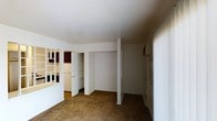 This single-floor space offers a straightforward and functional layout designed to maximize usability. The main living area features wall-to-wall carpeting in a warm neutral tone, providing a comfortable underfoot feel throughout. White walls and a clean, flat ceiling keep the space feeling bright and open.

A notable feature of the living area is the built-in shelving unit and closet storage, offering ample organizational capacity directly within the room. A structural column adds a subtle architectural element that helps define different zones within the open floor plan.

Adjacent to the main living area, a pass-through or open partition connects to the kitchen, which is visible with cabinetry in a darker finish and standard appliances, including a refrigerator. The kitchen layout appears compact yet efficiently arranged.

Natural light filters into the space through vertical blinds, softening the interior atmosphere. An additional door along one wall provides access to further areas or an exit point. The overall ambiance of the space is clean, neutral, and move-in ready, with practical storage solutions and a cohesive, understated design that suits a variety of uses.