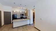This single-floor residence showcases a thoughtfully designed open-concept layout centered around a modern kitchen. The kitchen serves as the focal point of the living space, featuring granite countertops with an undermount sink and a peninsula that provides both additional workspace and a natural divider between the kitchen and adjoining areas. Light wood-tone cabinetry with clean-lined hardware extends to the ceiling, offering generous storage, while black appliances — including a refrigerator, microwave, and range — provide a striking contrast against the lighter finishes. Three pendant lights hang above the peninsula, complementing the recessed and track lighting that ensures the space is well-illuminated throughout. Light wood-look flooring runs continuously throughout the main area, contributing to a warm and cohesive aesthetic. The neutral white walls and clean architectural lines enhance the sense of openness. A hallway visible from the kitchen leads to additional rooms, including what appears to be a bathroom, suggesting a practical and efficient floor plan. A wall-mounted thermostat indicates individual climate control for added comfort.