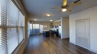 This single-floor space showcases a thoughtfully designed open-concept layout that seamlessly connects the living area and kitchen into one cohesive, airy environment. Rich, dark hardwood-style flooring flows throughout, grounding the space with warmth and contemporary style.

The kitchen is a standout feature, equipped with stainless steel appliances including a refrigerator, range, and microwave. Light wood cabinetry provides ample storage, while a dark-toned central island offers additional workspace and visual contrast. Pendant lighting hangs above the island, adding a refined touch to the culinary space.

The open living area benefits from generous natural light, courtesy of large windows fitted with horizontal blinds that allow for adjustable brightness. A ceiling fan with an integrated light fixture anchors the living space, ensuring both comfort and illumination.

The neutral wall tones and clean architectural lines contribute to a calm, modern ambiance throughout. The open sightlines from the living area to the kitchen make the space feel expansive and functional, ideal for both everyday living and entertaining. Overall, the design strikes a balance between practicality and contemporary aesthetic appeal.