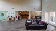 Located at 300 Caughman Farm Ln in Lexington, South Carolina, this single-floor community clubhouse offers a well-appointed gathering space designed for both socializing and resident services. The main lounge is the centerpiece of the facility, characterized by dramatically tall ceilings that amplify the sense of openness and light. A tile-surround fireplace anchors one end of the lounge, complemented by leather seating arranged on a patterned area rug over rich hardwood-style flooring. French doors lead directly to an outdoor area, allowing natural light to flow freely into the space. Adjacent to the lounge, a full kitchen is equipped with wood cabinetry and modern appliances, providing residents with a functional space for gatherings and events. A dedicated game room features a pool table, offering a recreational retreat. The leasing and administrative corridor is finished in a vibrant teal palette, lined with colorful artwork and recessed lighting, creating a bright and welcoming atmosphere. A small dining nook with a round wooden table and upholstered chairs provides an informal seating area near natural windows. Throughout, the consistent hardwood-style flooring, neutral wall tones, and curated décor lend the clubhouse a cohesive, modern aesthetic suited for community life.