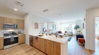 Located at 437 Cold Brook Dr in Sudbury, Massachusetts, this single-floor residence offers a thoughtfully designed open-concept layout with modern finishes throughout. The kitchen serves as a central hub of the home, featuring warm wood cabinetry, a large white quartz island with seating for multiple guests, stainless steel appliances, and a sleek linear pendant light overhead. Light hardwood flooring flows seamlessly from the kitchen into the living area, which is bathed in natural light from large windows overlooking lush greenery outside. The living room provides a comfortable and airy ambiance, with ample space for seating and relaxed gatherings. The bedrooms are generously sized, each offering large windows with serene outdoor views and light hardwood floors. One bedroom features a dedicated walk-in closet outfitted with wire shelving and upper storage. The bathroom is finished with a wood-toned vanity, white quartz countertop, a large mirror with modern lighting, and a tiled tub-shower combination. Throughout the residence, a neutral color palette of whites and warm wood tones creates a clean, contemporary aesthetic. The overall design balances functionality with style, making it an inviting and well-lit living space.