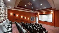 This dedicated screening room offers a true cinematic experience within a single-floor layout. The space is defined by its bold terracotta-colored walls, which contribute to the immersive, theater-like atmosphere. Rows of dark leather recliner-style seats with built-in cup holders are arranged in a tiered configuration, ensuring clear sightlines to the large projection screen mounted at the front of the room. A ceiling-mounted projector serves as the primary display source, suspended from a tray ceiling detail that adds architectural interest overhead. Recessed lighting is integrated into the ceiling, complemented by a series of elegant wall sconces that provide warm, ambient illumination along the side walls. Decorative abstract artwork is displayed on one wall, adding a refined touch to the entertainment-focused interior. The patterned carpet underfoot adds both comfort and visual texture to the space. Wall-mounted speakers are positioned throughout to support a full audio experience. The overall design balances functionality and comfort, creating an inviting and well-appointed venue suited for private screenings and group viewing.