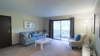 This single-floor residence offers a comfortable and well-designed living space with a clean, functional layout. The living room serves as the centerpiece of the home, featuring plush wall-to-wall carpeting that adds warmth underfoot and a soft, neutral color palette throughout. Large sliding glass doors dominate one wall, flooding the room with natural light and providing direct access to an outdoor area with greenery visible beyond. Charcoal curtain panels frame the doors, offering both style and privacy when needed. The living area is furnished with a blue sofa accented by patterned throw pillows, a sleek white coffee table, and a coordinating accent chair, creating a cohesive and inviting atmosphere. A framed piece of wall art adds a finishing touch to the understated décor. The muted tones of the walls and ceiling complement the overall calm and airy ambiance of the space. The single-floor layout ensures ease of movement and accessibility throughout. Overall, this residence balances practicality with a relaxed, welcoming aesthetic, making it a comfortable everyday living environment enhanced by its connection to natural outdoor surroundings.