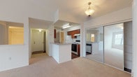 This single-story residence is designed with an open, functional layout that connects the main living spaces seamlessly. The living area is a standout feature, distinguished by vaulted ceilings that lend a sense of openness and volume to the space. Soft wall-to-wall carpeting runs throughout the living and bedroom areas, contributing to a warm, comfortable atmosphere. A ceiling fan with integrated lighting provides both airflow and illumination, while a pendant light fixture accents the entry to the kitchen area. Mirrored sliding closet doors along one wall of the living space reflect light and add visual depth to the room.

The kitchen is configured in a galley style, featuring rich wood-toned cabinetry, light countertops, a double-basin sink, a gas range, a white refrigerator, and a dishwasher. Dark hardwood-style flooring runs throughout the kitchen, offering a practical contrast to the carpeted areas. A peninsula counter connects the kitchen to the living space, creating a natural boundary while maintaining an open feel.

The bedroom is generously sized, carpeted, and includes its own ceiling fan. A pass-through connects it to the kitchen area, enhancing the unit's functional flow. The bathroom features wood-style flooring, a rich vanity cabinet, and a full tub with tiled surround and overhead shower.