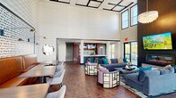 Located at 2541 Landmark Dr in Raleigh, NC, this residential community amenity space delivers an upscale, resort-inspired experience across thoughtfully designed indoor and outdoor areas. The centerpiece of the interior is a dramatic double-height clubroom bathed in natural light from expansive floor-to-ceiling windows. A coffered ceiling, gleaming chandelier, and linear fireplace set an elegant tone, while plush sectional sofas, accent chairs, and rich dark hardwood-style flooring create a welcoming social atmosphere. An adjacent lounge and dining area features leather banquette seating against a striking geometric accent wall, complemented by stylish pendant lighting. A well-appointed kitchenette with sage subway tile backsplash, quartz countertops, and warm cabinetry provides a convenient refreshment station for residents. Outdoors, a generously sized resort-style pool serves as the community's recreational focal point, surrounded by ample lounge seating and a broad paver deck. A landscaped courtyard offers a pergola-covered gathering space with seating, manicured greenery, and decorative lighting, ideal for outdoor relaxation. The multi-building community features modern architectural exteriors blending brick, panel, and glass elements, with private balconies overlooking the grounds.
