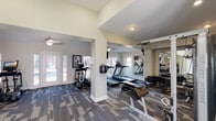 This fitness center is thoughtfully designed across a single floor, offering a generous and open layout divided into several interconnected workout zones. The space features varying ceiling heights throughout, with soaring vaulted and elevated ceilings in certain sections that contribute to an airy, expansive atmosphere. Ceiling fans are positioned throughout to maintain airflow and comfort during workouts.

The cardio zone is anchored by a row of treadmills and elliptical machines, positioned directly in front of large windows and glass doors that overlook an outdoor pool area, providing motivating views and an abundance of natural light. Wall-mounted televisions are conveniently placed throughout the space for entertainment during exercise.

The strength training area includes a multi-station cable machine, free weights, adjustable benches, and additional resistance equipment. Full-length mirrors line the walls in this section, visually expanding the space and allowing for proper form monitoring. Motivational wall art adds a welcoming, energetic character to the environment.

The flooring throughout consists of durable, patterned carpet tiles in a neutral gray tone, providing cushioning underfoot. Multiple exterior doors offer easy access and enhance ventilation, while the light green and white walls maintain a clean, refreshing aesthetic throughout the entire facility.