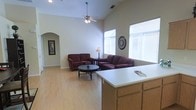 This single-story home showcases an open-concept design that seamlessly connects the kitchen and living areas. The living room is defined by its vaulted ceilings, which add a sense of spaciousness and volume to the space, while a ceiling fan with a light fixture provides both comfort and illumination. Large windows with white blinds allow ample natural light to fill the room, creating a bright and welcoming atmosphere.

The flooring throughout is light-toned wood, lending warmth and continuity to the open layout. An arched doorway on one wall adds an architectural detail that introduces a touch of elegance while providing a visual transition to adjacent areas of the home.

The kitchen is partially visible in the foreground, featuring light oak cabinetry and a white countertop that extends into a peninsula or breakfast bar, serving as a natural divider between the kitchen and living spaces. This layout facilitates easy interaction between the two areas, making it well-suited for everyday living and entertaining. The neutral color palette of beige and white walls throughout contributes to a clean, airy aesthetic.