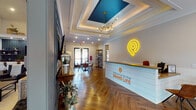 The Grand Life Clubhouse is a thoughtfully designed single-floor community space that blends modern aesthetics with warm, inviting details throughout. Guests are welcomed at a crisp white reception desk accented with the "Welcome to the Grand Life" branding and a glowing neon logo, all set beneath a stunning coffered ceiling with a bold teal inset and a sculptural brass chandelier. Herringbone-patterned hardwood flooring runs seamlessly throughout the entire space, grounding each area with a consistent sense of refined style. The main lounge area features soaring vaulted ceilings with exposed dark wood beams, deep teal accent walls, and a wall-mounted flat-screen television framed by decorative molding above a rustic wooden credenza. Comfortable leather seating and layered textiles create an inviting atmosphere ideal for relaxing or socializing. Adjacent to the lounge, a dining area is bathed in natural light streaming through large sliding glass doors that open to an outdoor patio view. A substantial round wooden dining table surrounded by leather chairs anchors the room, complemented by open shelving units and patterned drapery. The wet bar area is a standout feature, showcasing a dramatic dark navy subway tile backsplash, under-counter beverage refrigerators, a stainless steel sink, floating wooden shelves, and a generous island with tufted white bar seating — perfect for entertaining.