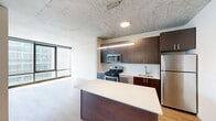 Located in Chicago's vibrant South Loop neighborhood, this modern one-bedroom, one-bathroom unit offers a well-designed open-concept layout with a seamless flow between the kitchen, living, and dining areas. The space is defined by striking exposed concrete ceilings throughout, warm light wood flooring, and expansive floor-to-ceiling windows that flood the interior with natural light and frame city views.

The kitchen is thoughtfully appointed with rich dark-walnut cabinetry, a sleek white quartz waterfall island, and a full suite of stainless steel appliances including a gas range, microwave, dishwasher, and refrigerator. A pendant linear light fixture over the island adds a contemporary touch.

The open living area benefits from generous natural light through large windows overlooking the Chicago skyline, creating an airy, inviting atmosphere. A dedicated hallway provides access to the bedroom and reveals a discreetly tucked-away in-unit stacked washer and dryer — a rare and practical convenience.

The bedroom is a bright, calm retreat with its own floor-to-ceiling corner windows offering sweeping urban views. The bathroom is finished in a clean, modern style with large-format marble-look tiles in the walk-in glass shower, a wood-toned vanity, and light wood flooring that ties the space together cohesively.