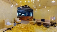 Scout on the Circle is a boldly designed, single-floor communal space that makes an immediate impression upon entry. The entrance features floor-to-ceiling glass doors framed in dark steel, flanked by warm wood paneling and polished concrete flooring, establishing a modern industrial aesthetic from the outset.

The primary lounge zone is anchored by a striking golden-yellow patterned carpet and textured 3D wall panels in matching tones. A dramatic chandelier installation of exposed Edison bulbs suspended on angular wire forms cascades from a yellow geometric ceiling feature, creating a warm, artistic focal point. Modular sectional seating and egg-style lounge chairs furnish this area generously.

A tiered wooden platform provides a casual, amphitheater-like gathering space that transitions into a darker-toned back lounge. This area features dark wood-paneled walls, built-in bench seating with colorful upholstery, a wall-mounted display, and windows overlooking an outdoor area.

A dedicated meeting nook includes an oval dark conference table, wire-frame chairs, a vertical slatted partition, and a multicolored sculptural chandelier overhead. Hardwood flooring grounds this zone against the surrounding concrete.

Throughout, exposed ductwork and open ceilings reinforce the industrial design language, while curated artwork, bold color-blocking, and varied textures give the space a dynamic, creative character suited for collaboration and community engagement.