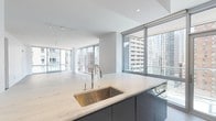 This contemporary high-rise residence is defined by a sleek, modern aesthetic and an abundance of natural light. The open-plan living and kitchen area serves as the centerpiece of the space, anchored by a substantial island with elegant marble countertops and a stainless steel undermount sink. The kitchen is fitted with high-gloss gray cabinetry, a marble slab backsplash, and a professional-grade stainless steel range with a gas cooktop. Light hardwood flooring flows through the main living areas, complementing the clean white walls and minimalist design sensibility. Floor-to-ceiling windows and glass doors frame sweeping urban skyline views throughout, flooding the living space with natural light. The bedrooms feature plush neutral carpeting and large windows that continue the connection to the cityscape. Ample built-in closet storage is present in the sleeping quarters. The bathrooms are finished with crisp white subway tile, soaking tubs with glass shower enclosures, chrome fixtures, and warm wood-toned vanities set against light tile flooring. Track lighting in the living area and recessed fixtures throughout provide versatile illumination. The overall ambiance is polished, bright, and contemporary, offering a refined urban living experience.