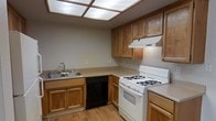 This single-floor residence presents a well-organized layout with distinct living zones connected by a central corridor. The kitchen is a standout functional area, equipped with warm honey-toned wood cabinetry offering ample upper and lower storage. It features a double-basin stainless steel sink, a white gas range with a ventilation hood, a black dishwasher, and continuous countertops that provide generous prep space. A luminous drop ceiling with panel lighting brightens the kitchen, complemented by light hardwood-style flooring that adds warmth underfoot. Adjacent living areas feature plush wall-to-wall carpeting in a neutral tone, creating a comfortable and cohesive atmosphere. One of the larger rooms benefits from vaulted or elevated ceilings, adding a sense of openness and volume to the space. Vertical blinds cover a sliding door or wide window, allowing natural light to filter in while offering privacy. The overall color palette throughout is neutral, with light gray and beige walls that provide a clean, versatile backdrop. The floor plan, as seen in the dollhouse view, reveals a thoughtfully segmented layout with multiple rooms suitable for varied uses, making this a practical and adaptable living space.