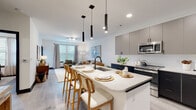 This thoughtfully designed single-floor residence offers a seamless blend of modern aesthetics and functional living. The open-concept kitchen and living area serves as the heart of the home, anchored by a large center island with quartz countertops and seating for multiple guests, illuminated by sleek pendant lighting above. The kitchen is outfitted with two-tone cabinetry — pairing dark lower cabinets with warm gray uppers — complemented by a white subway tile backsplash and a full suite of stainless steel appliances, including a refrigerator, range, and over-the-range microwave. Light wood-look flooring flows throughout, tying the spaces together cohesively. The living area is bright and inviting, with large windows that allow generous natural light and views of greenery beyond. The bedroom is a calm retreat, featuring the same wood-look flooring, a ceiling fan, and wide windows dressed with linen curtains that enhance the airy atmosphere. The bathroom is generously proportioned, finished with large-format tile flooring, a glass-enclosed shower, a vanity with storage, and direct access to a dedicated walk-in closet. An in-unit stacked washer and dryer rounds out the practical amenities of this well-appointed residence.