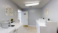 Located at 4426 Calderwood Drive in Columbus, Ohio, this single-floor laundry facility is designed for practical, efficient use. The space is outfitted with multiple commercial-style washing machines and dryers arranged along the walls, providing ample capacity for residents or users. The room features light-colored tile flooring that is easy to maintain and contributes to a clean, functional atmosphere. Soft blue-gray walls give the space a calm and neutral feel, while bright fluorescent overhead lighting ensures excellent visibility throughout. A white-paneled door with a gold handle leads to an exterior entry point, allowing for convenient access and natural light to filter in. Ventilation is provided via ceiling vents, supporting a comfortable environment during use. Wall-mounted signage, including a clearly labeled "Laundry" designation, helps orient users within the space. The overall layout is straightforward and well-organized, with cabinetry providing additional storage and surface space. This facility offers a clean, well-lit, and accessible environment suited to everyday laundry needs.