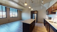 This single-floor urban loft showcases a compelling blend of industrial and modern design. The open-plan living and kitchen area is defined by soaring ceilings with exposed concrete and visible ductwork, giving the space a distinctive warehouse-inspired character. Pendant and track lighting illuminate the area, highlighting the warm-toned hardwood flooring that runs throughout.

The kitchen is a standout feature, equipped with rich dark wood cabinetry, stainless steel appliances including a refrigerator, range, and microwave, and a generous center island with a clean white countertop that doubles as a prep and dining surface. A galley-style layout provides ample countertop and storage space along one wall, complemented by a stainless steel sink.

Large industrial-style windows flood the living area with natural light while offering sweeping city skyline views, reinforcing the urban loft aesthetic. The open living space is expansive and flexible, well-suited to a variety of furniture arrangements.

The bathroom is finished to a clean, contemporary standard, featuring large-format tile flooring, a glass-enclosed walk-in shower with textured tile surround, a dual-sink vanity with dark cabinetry, and neutral wall tones that create a calm, functional atmosphere. Overall, this space delivers a cohesive industrial-modern aesthetic with quality finishes throughout.