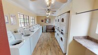 This shared laundry facility is thoughtfully designed to accommodate a high volume of users in a comfortable, functional environment. The single-floor space is finished with warm wood-look flooring and soft yellow walls trimmed with white crown molding, creating a clean and welcoming atmosphere.

A long row of top-load washing machines lines one wall, each equipped with digital controls for user convenience. Opposite the washers, multiple stacked dryer units maximize the available space while providing ample drying capacity. A built-in laminate folding counter runs along one wall beneath large, grid-paned windows that invite natural light and offer views of the surrounding property.

Ceiling fans with light fixtures are mounted throughout the space, ensuring comfortable air circulation during use. A wood-slat bench provides a comfortable waiting area, while a cork bulletin board displays posted notices and community information. A Coca-Cola vending machine near the entrance adds an extra layer of convenience for users.

The facility also includes a paper towel dispenser and a waste receptacle, reflecting attention to cleanliness and practicality. Overall, the space balances utility and comfort, offering residents a well-maintained and efficiently organized laundry experience.