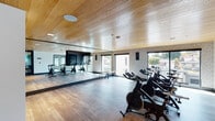 Located at 4949 Baltimore Dr in La Mesa, California, this contemporary fitness studio is thoughtfully designed to deliver an energizing and functional workout environment. The space is defined by its striking warm wood-panel ceiling, which lends a natural, upscale aesthetic throughout. The primary workout area features a dedicated spin cycling room equipped with multiple stationary bikes arranged to face large picture windows that frame views of the surrounding neighborhood, flooding the space with generous natural light. Full-length mirrored walls run the length of the room, enhancing the sense of openness and allowing exercisers to monitor their form during sessions. A geometric-patterned accent wall adds a modern, design-forward touch to the interior. Adjacent to the cycling area, a well-stocked fitness zone houses an assortment of exercise equipment including stability balls, medicine balls, resistance bands, yoga mats, and balance tools organized on dedicated storage racks. Sleek black-framed glass doors connect the individual workout zones, maintaining visual continuity while providing separation between spaces. The durable hardwood-style flooring ties the entire facility together, complementing the clean, contemporary design. The overall ambiance is bright, airy, and motivating, making this fitness studio a premium amenity for the La Mesa community.