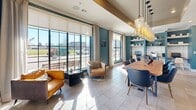 This well-appointed single-floor space seamlessly combines a dining area and living room into one cohesive, open-plan environment. The interior is defined by striking teal accent walls that contrast elegantly against crisp white paneling and light-toned tile flooring, creating a modern yet inviting atmosphere throughout.

At the heart of the dining area sits a substantial live-edge wood communal table surrounded by contemporary navy blue chairs, anchored overhead by a dramatic cascading cluster of amber glass pendant lights. This eye-catching fixture serves as the focal point of the room and adds a warm, sculptural quality to the space.

The living area features a pair of caramel leather armchairs arranged to take full advantage of the floor-to-ceiling black-framed glass walls and large multi-pane windows, which flood the interior with abundant natural light and offer open views of the outdoors. Sheer linen curtains frame the windows, softening the light while maintaining an airy feel.

A built-in workstation is tucked along one teal wall, complete with open wood shelving for display and storage, alongside a countertop workspace. A white board-and-batten feature wall houses a wall-mounted display screen, adding a functional yet polished finishing touch. The space is cohesive, purposeful, and thoughtfully designed for both collaboration and relaxation.