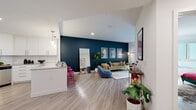 Located at 5401 Lankershim Blvd in North Hollywood, California, this stylish single-floor residence offers a thoughtfully designed open-concept layout that seamlessly connects the living, dining, and kitchen areas. The living room is a striking focal point, anchored by a deep teal accent wall adorned with a curated gallery of vibrant artwork. Large sliding glass doors flood the space with natural light and open to a private balcony, enhancing the sense of openness. Light wood-style flooring runs throughout, providing a warm contrast to the bold color palette. The kitchen is outfitted with crisp white cabinetry, a classic subway tile backsplash, quartz countertops, stainless steel appliances — including a gas range and dishwasher — and a peninsula with pendant lighting that doubles as a casual dining counter with colorful bar seating. The bathroom features a clean, contemporary design with a full soaking tub and shower combination, wood-toned vanity cabinetry, and white quartz countertops. The entryway is practical and well-organized, offering louvered closet doors and convenient access to in-unit storage. Throughout the residence, recessed lighting and high ceilings contribute to an airy, modern ambiance ideal for comfortable urban living in the heart of North Hollywood.