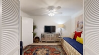 This single-story home accommodates five bedrooms within a thoughtfully organized floor plan. The living room serves as the centerpiece of the home, featuring a classic fireplace flanked by tall indoor plants, large windows with horizontal blinds that invite natural light, and carpeted flooring. A built-in shelving nook adjacent to the living area provides practical storage and display space. The dining room is compact and intimate, highlighted by a round glass-top table and a decorative brick accent wall, complemented by a striking sunburst mirror. The kitchen is fitted with white cabinetry, light stone countertops, a subway tile backsplash, tile flooring, and black appliances including a refrigerator, dishwasher, and electric range. The shared bathroom features a wide vanity with stone-look countertops, white cabinetry, and a large mirror. The primary bedroom is generously sized, featuring a ceiling fan, a window with outdoor views, and plush carpeting. The additional bedrooms are bright and well-proportioned, each with ceiling fans, windows overlooking greenery, and carpeted floors. A carpeted hallway connects the bedrooms and shared spaces efficiently, maintaining a clean and neutral aesthetic throughout the home.