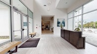 This single-story clubhouse and leasing facility presents a bright, contemporary design characterized by soaring ceilings, light-toned wood-look tile flooring throughout, and an abundance of natural light. The main entrance welcomes visitors through glass double doors, framed by transom windows that flood the foyer with daylight. A tufted bench provides convenient seating near the entry, while a sleek dark wood reception desk with a light stone countertop anchors the front area. Floor-to-ceiling windows behind the reception desk offer direct views of an outdoor pool area, creating a seamless indoor-outdoor connection.

Adjacent to the lobby, a well-appointed lounge area features stylish swivel chairs, a round coffee table, and an artistic pendant light fixture, with additional windows overlooking the pool. A dedicated dining and café area includes built-in banquette seating, bistro tables, and elegant chandelier lighting beneath a coffered ceiling, accompanied by a full Starbucks coffee station with a large island and bar seating. Geometric decorative panels and open shelving with a seafoam tile backsplash add visual interest to the kitchen zone. A spacious game room rounds out the amenities, featuring a billiards table, foosball, a teal accent wall with decorative art, and ample seating.