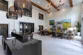 Located at 659 Treeland Dr in Ladson, South Carolina, this single-story community space is designed with both functionality and aesthetic appeal in mind. The layout is open and expansive, anchored by dramatically high ceilings that amplify the sense of volume throughout the interior.

The entry area features bold black-framed double doors flanked by full-length sidelights, allowing natural light to flow generously into the space. Light-toned tile flooring laid in a herringbone pattern extends throughout, providing a clean, modern foundation that unifies each area.

The lounge area serves as the visual centerpiece of the space. A decorative wall with a paneled grid detail acts as a gallery backdrop for an eclectic collection of colorful artwork. Comfortable seating arrangements—including dark upholstered sofas and accent chairs with vibrant throw pillows—are anchored by rich wood coffee tables and layered area rugs, creating an inviting and well-curated atmosphere.

Floor-to-ceiling windows dressed with patterned drapery introduce additional natural light and outdoor views into the lounge. The kitchenette area features dark cabinetry, a wall-mounted television, shiplap-style wall accents, pendant lighting, and beverage station appliances, offering practical amenities in a stylish setting.