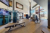 This modern fitness center is defined by its dramatic double-height ceilings and expansive floor-to-ceiling grid-pane windows that flood the space with natural light and provide open views of the surrounding landscape. A sculptural spiral staircase serves as a standout architectural focal point, rising elegantly from the main floor to an upper mezzanine level with metal railings overlooking the gym below.

The fitness floor is thoughtfully organized into dedicated zones. The cardio area features multiple Matrix treadmills and elliptical trainers positioned directly in front of the large windows, creating an energizing workout environment. The strength training section is comprehensively outfitted with Matrix cable machines, multi-functional weight benches, a full rack of dumbbells, and a variety of selectorized resistance machines. A dedicated functional training zone includes a RAX multi-tier storage rack stocked with colorful kettlebells, medicine balls, and slam balls, complemented by TRX suspension trainers.

The space is finished with durable rubber-blend flooring throughout, dark accent columns, and strategically placed mirrors that enhance the sense of space. Recessed lighting and a mounted flat-screen television complete the polished, professional ambiance of this fully appointed fitness facility.
