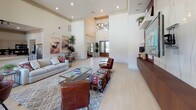 This sophisticated single-floor community space is defined by its dramatic double-height ceilings and an open, airy layout that creates an immediate sense of grandeur. Light-toned tile flooring runs throughout, complementing the bright, neutral wall palette and recessed lighting that keeps the interior feeling inviting and well-illuminated.

The main lounge area serves as the centerpiece of the space, anchored by a striking feature wall clad in veined marble-style stone. Dark wood built-in shelving flanks the wall-mounted television, providing display space for curated décor. Leather accent chairs in rich cognac tones, a patterned ottoman, and a glass-topped coffee table are arranged over a geometric area rug, creating a comfortable and visually engaging seating zone.

Adjacent to the lounge, a dining area with a dark table and leather chairs is positioned near floor-to-ceiling windows and a glass door that opens to views of a pool area beyond, flooding the space with natural light. Abstract artwork adorns the walls, adding color and personality throughout.

A café-style counter with tall seating provides a casual gathering spot, while an open catering kitchen is visible in the background. A dedicated workstation or business center alcove is tucked to one side, offering a quiet area for productivity. Tall tropical potted plants punctuate the space, adding organic warmth to the polished interior design.