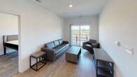 This single-floor unit offers a clean, contemporary design throughout. The kitchen is well-appointed with dark espresso cabinetry, granite countertops, a stainless steel sink integrated into a peninsula, and a full suite of stainless steel appliances including a refrigerator, electric range, and microwave. White subway tile backsplash adds a polished touch, while light wood-style flooring ties the space together.

The living area features the same light wood-style flooring and is furnished with dark leather seating. A sliding glass door opens to a private balcony, allowing natural light to fill the room and providing views of the surrounding exterior.

Multiple bedrooms are present, each carpeted and equipped with a metal-frame bed and wood-toned dresser. The bedrooms include windows that bring in natural light and offer exterior views. One bedroom features a connected en-suite bathroom, enhancing privacy and convenience.

The bathrooms are finished consistently with dark vanity cabinetry, granite countertops, and light wood-style flooring. One bathroom includes a bathtub/shower combination, while the other serves as a more compact layout. Brushed nickel fixtures and hardware complete the refined, cohesive aesthetic found throughout the unit.