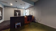 This single-floor indoor sports simulator facility is purpose-built for golf and multi-sport simulation, offering an immersive and functional environment for players of all skill levels. The space centers around a TruGolf simulator bay, enclosed by dark surround walls that enhance projection quality and focus attention on the large high-definition screen. The simulator supports a variety of experiences, including virtual golf courses and sports games such as Breakaway Soccer, making it a versatile entertainment and training destination.

The reception and staging area is finished with neutral gray walls, green turf-style flooring, and sleek pendant lighting that creates a modern, sports-oriented ambiance. A dark front counter is complemented by a warm wood storage unit housing multiple golf bags and equipment, keeping the space organized and ready for use. Bold black-and-white silhouette artwork depicting golfers and athletes lines the walls, reinforcing the sporty character of the venue.

A dedicated control station with a wall-mounted monitor and wooden cabinet provides easy access to simulator settings and game selection. The turf flooring throughout ties the interior aesthetic together, evoking the feel of a real course. The overall design is sleek, dark-toned, and intentional, delivering a professional atmosphere suited for both casual play and serious practice sessions.