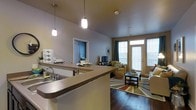 This single-floor residence offers a thoughtfully designed open-concept layout that seamlessly connects the kitchen and living areas. The kitchen is anchored by a generously sized breakfast bar, complete with an integrated sink and pendant lighting that hangs from the ceiling, providing both task illumination and a stylish focal point. The bar extends to offer casual seating and dining space, making it ideal for everyday use and entertaining.

The living area is bathed in natural light streaming through large windows and a set of glass-paneled doors that lead to a private balcony, creating a bright and airy atmosphere. Rich hardwood flooring runs throughout, adding warmth and a sense of continuity to the open floor plan. The living space comfortably accommodates a full seating arrangement, centered around a coffee table and complemented by a colorful area rug that adds a vibrant touch to the interior.

Neutral-toned walls and smooth ceilings throughout the space create a clean, cohesive backdrop that allows furnishings and natural light to take center stage. A doorway visible from the main living area hints at additional rooms beyond, rounding out this well-appointed and functional single-floor home.