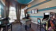 This single-floor amenity space is designed as an engaging recreational and social hub, combining a dedicated game room with adjoining lounge and kitchen areas. The game room is a standout feature, outfitted with a full-size foosball table, a multi-game arcade cabinet, and a classic pinball machine themed around The Munsters. The space is anchored by a distinctive design palette, with bold teal horizontal accent stripes running along the walls, a teal-painted tray ceiling, and a sculptural pendant light fixture that adds a retro-modern flair. Tall windows dressed in bold, patterned drapery panels flood the room with natural light while providing visual interest. Patterned carpet tiles in brown and gray tones ground the space and complement the overall color scheme. A wall-mounted flat-screen television adds to the entertainment options. Glass partition walls with a sliding entry door separate the game room from an adjacent area featuring high-top bistro tables and bar-height seating. Through an open doorway, a well-appointed kitchen with cabinetry is visible, supporting the space's function as a community gathering area. The overall ambiance is energetic, colorful, and thoughtfully designed for leisure and social interaction.