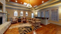 Located at 3374 Starboard Way in Fayetteville, NC, this single-floor community clubhouse offers a well-appointed gathering space designed for comfort and functionality. The main lounge area is a standout feature, showcasing rich hardwood flooring, a dramatic tray ceiling with a bold burnt-orange inset and elegant chandelier, and detailed crown molding throughout. Wall sconces illuminate a curated gallery of framed artwork, while generous seating — including plush upholstered chairs and a full sofa — creates a welcoming atmosphere for socializing. An adjacent kitchenette is outfitted with rich wood cabinetry, granite-style countertops, a peninsula with bar seating, a stainless steel refrigerator, a sink, and decorative pendant lighting, making it ideal for hosting events. The ADA-accessible restroom features tile wainscoting, granite countertops, and safety grab bars, reflecting thoughtful, inclusive design. A spacious covered outdoor patio with classic brick pavers, a ceiling fan, and stone-accented exterior walls provides a comfortable transition to the outdoor pool area, complete with a water fountain station and gated access. Natural light flows throughout via multiple windows, enhancing the warm, inviting ambiance of this versatile community space.