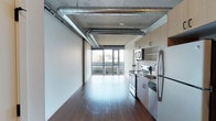 Located at 624 Yale Ave N in Seattle's vibrant South Lake Union neighborhood, this modern urban residence showcases an industrial-chic aesthetic that blends raw architectural elements with contemporary finishes. The open-concept living area is defined by soaring exposed concrete ceilings with prominent silver ventilation ductwork, lending an authentic loft-like character to the space. Rich, dark hardwood floors run throughout, providing a warm contrast to the cool industrial palette above. Floor-to-ceiling windows along the exterior wall flood the interior with abundant natural light while framing sweeping views of the Seattle skyline — a standout feature that brings the energy of the city directly into the home. The kitchen is positioned along one side of the main living area and is equipped with stainless steel appliances, including a refrigerator, electric range, and built-in microwave. Light wood cabinetry provides a clean, modern storage solution, complementing the open layout seamlessly. The overall ambiance is sleek and metropolitan, combining the utilitarian charm of exposed building systems with refined materials and finishes. This single-floor residence offers a compelling urban living experience in one of Seattle's most sought-after and walkable neighborhoods.