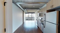 Located at 624 Yale Ave N in Seattle's vibrant South Lake Union neighborhood, this modern urban residence showcases an industrial-chic aesthetic that blends raw architectural elements with contemporary finishes. The open-concept living area is defined by soaring exposed concrete ceilings with prominent silver ventilation ductwork, lending an authentic loft-like character to the space. Rich, dark hardwood floors run throughout, providing a warm contrast to the cool industrial palette above. Floor-to-ceiling windows along the exterior wall flood the interior with abundant natural light while framing sweeping views of the Seattle skyline — a standout feature that brings the energy of the city directly into the home. The kitchen is positioned along one side of the main living area and is equipped with stainless steel appliances, including a refrigerator, electric range, and built-in microwave. Light wood cabinetry provides a clean, modern storage solution, complementing the open layout seamlessly. The overall ambiance is sleek and metropolitan, combining the utilitarian charm of exposed building systems with refined materials and finishes. This single-floor residence offers a compelling urban living experience in one of Seattle's most sought-after and walkable neighborhoods.