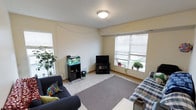 This single-floor residential space offers a comfortable and functional layout centered around a warm, inviting living area. The room is finished with light yellow-toned walls and a neutral tile floor, partially covered by a gray area rug that helps define the seating zone. A flush-mount ceiling light provides overhead illumination, complementing the abundant natural light that streams in through multiple windows fitted with white horizontal blinds. The living area accommodates a pair of sofas — a dark navy blue couch with colorful accent pillows and a classic blue-and-white plaid upholstered sofa — arranged to create a relaxed, conversational layout. A flat-screen television sits atop a black media console, serving as the focal point of the entertainment area. Adding a unique and lively touch to the space is a fish aquarium displayed on a dedicated stand, bringing color and movement to the room. Potted plants positioned near the windows further enhance the natural, homey feel. The overall ambiance is casual and comfortable, with personal touches that give the space a distinctly lived-in character. The single-floor layout ensures easy accessibility and a cohesive flow throughout.