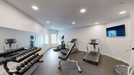 Located at 2274 5th Ave in Marion, Iowa, this dedicated exercise room offers a well-equipped fitness space designed to support a comprehensive workout routine. The room is finished with clean white walls and durable rubber flooring throughout, providing both a functional and tidy environment for exercise.

The space is outfitted with multiple cardio machines, including treadmills and elliptical trainers, allowing users to engage in sustained aerobic activity. A stationary air bike adds an additional option for high-intensity interval training. A padded adjustable weight bench and a rack of dumbbells in varying weights support strength and resistance training. A wall-mounted flat-screen television provides entertainment during workouts.

A large wall mirror spans a significant portion of one wall, enhancing the sense of space and allowing users to monitor their form. Natural light filters in through a window with blinds, complementing the recessed ceiling lighting that keeps the room well-lit at all times. The fitness center is accessible through a set of double doors, offering easy entry and exit. The overall atmosphere is clean, practical, and purpose-built, making this exercise room a convenient and fully functional amenity for fitness-focused individuals.