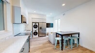 Located at 2137 Dwight Way in Berkeley, CA, this 5-bedroom, 2-bathroom unit offers a thoughtfully designed layout across a single floor. The open-concept kitchen and living area serves as the heart of the unit, featuring two-tone cabinetry in teal and light gray, stainless steel appliances including a range, dishwasher, microwave, and French door refrigerator, plus a stacked in-unit washer and dryer. Light wood-look flooring runs throughout, contributing to a clean, contemporary aesthetic. A dining bar with stools provides casual seating alongside a couch, making the shared living space both functional and welcoming. The five bedrooms are compact and efficiently designed, each featuring light wood bed frames, windows that bring in natural light, and views of the surrounding Berkeley neighborhood. Some bedrooms also include a desk and chair, offering a dedicated workspace. The two bathrooms are finished consistently with teal vanity cabinetry, marble-style countertops, black-framed mirrors, and wood-look flooring. One bathroom features a tub-shower combo, while the other offers a shower with a streamlined layout. Recessed lighting throughout the unit adds a modern touch, and the overall palette of white walls and neutral tones creates a bright, cohesive living environment well-suited for shared occupancy.