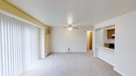 This single-floor space centers around an open living area finished with light-toned wall-to-wall carpeting and white walls that create a bright, clean atmosphere. A ceiling fan with an integrated light fixture is mounted overhead, providing both air circulation and ambient lighting. Along one wall, a wall-mounted air conditioning unit offers supplemental climate control, complemented by a baseboard heating unit positioned along the perimeter for year-round comfort. Vertical blinds frame a large window or sliding door opening on the left side of the room, allowing natural light to filter in while offering privacy options. The layout opens toward additional rooms in the background, where a warm-toned wooden door is visible, hinting at a private room beyond. To the right, a partial wall with a pass-through counter suggests a connected kitchen area, adding functional convenience to the open floor plan. The overall design is straightforward and practical, with neutral finishes throughout that provide a versatile canvas for personalization. The single-floor configuration ensures easy accessibility and a cohesive flow between living spaces.