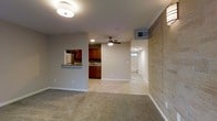 This single-floor space is defined by an open-concept layout that seamlessly connects the living, dining, and kitchen areas. The living area is finished with plush carpeting, while the kitchen and adjacent spaces transition to light-tiled flooring, creating a clear visual distinction between zones. A prominent stone tile accent wall anchors one side of the living area, adding texture and a refined architectural element to the interior.

The kitchen features warm-toned wood cabinetry, a countertop with a pass-through window opening into the living area, and a sink positioned along the back wall — a practical and social design choice. Recessed lighting and a ceiling fan with a light kit serve the main open area, ensuring both comfort and functionality.

A hallway visible from the main living space leads toward additional rooms, with natural light visible at the far end suggesting a window or exterior-facing room. HVAC venting is integrated into the ceiling, and the neutral gray wall tones throughout provide a clean, versatile backdrop. The overall ambiance is bright, practical, and well-organized.