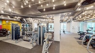 This expansive fitness center offers a comprehensive workout environment designed to accommodate a wide range of exercise disciplines. The space features an industrial-chic aesthetic defined by exposed ductwork, structural columns, and an eye-catching array of Edison bulb pendants suspended from the ceiling, creating a warm, energetic ambiance throughout.

The main floor is thoughtfully organized into distinct workout zones. A dedicated strength training area houses a full lineup of Matrix cable and selectorized machines, along with multiple squat racks, barbells, and weight plates positioned on padded floor mats. A free weights section provides dumbbells and functional training accessories, while a vibrant, color-blocked mural adds a bold artistic focal point to this zone.

The cardio area features a row of Matrix treadmills and other aerobic equipment positioned near floor-to-ceiling windows, allowing natural light to fill the space and offering outdoor views. A separate, glass-enclosed spin studio with bold yellow accent walls, cycling bikes, and a mounted display screen provides a dedicated space for group or individual cycling sessions. Striped carpet tiles unify the flooring throughout, contributing to a cohesive, modern design.