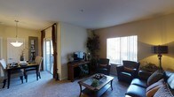 This single-floor residence offers a thoughtfully arranged living space designed for both comfort and functionality. The living room serves as the heart of the home, featuring plush wall-to-wall carpeting that adds warmth underfoot. Large windows fitted with vertical blinds fill the room with generous natural light, while a sliding glass door provides seamless access to an outdoor patio area, enhancing the sense of openness and connection to the exterior. The living room is furnished with a rich dark leather sofa and coordinating armchairs, centered around a classic wood-and-glass coffee table that anchors the seating arrangement. A wood media console houses a flat-screen television, complemented by decorative accents including tall candleholders and a lush potted indoor tree that adds a natural, organic element to the space. Neutral-toned walls and a smooth white ceiling create a clean, airy backdrop that allows the dark wood and leather furnishings to stand out. Floor-length drapes frame the patio entrance, adding a layer of elegance and privacy. Overall, the space presents a warm and inviting ambiance, well-suited for relaxed everyday living and casual entertaining.