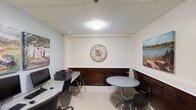This single-floor workspace is designed to support both focused productivity and informal collaboration. The room features a clean, neutral color palette with white walls and a drop-panel ceiling equipped with flush-mounted overhead lighting, creating a bright and functional atmosphere. Dark wood wainscoting lines the lower portion of the walls, adding a touch of warmth and a classic aesthetic contrast to the otherwise modern setup. A large decorative wall clock serves as a central focal point, complementing a pair of landscape art pieces that add color and visual interest to the space. On one side of the room, a dedicated computer workstation accommodates a desktop monitor, printer, and ergonomic seating, making it well-suited for tasks that require technology access. On the opposite side, a round table with chairs provides a convenient spot for informal meetings, light reading, or casual work. The light-toned carpet underfoot contributes to the room's inviting and comfortable feel. Overall, this space strikes a balance between professional functionality and a welcoming ambiance, making it a practical resource for anyone in need of a quiet, equipped environment to work or collaborate.