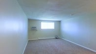 This single-floor space offers a straightforward and functional layout designed for comfortable living. The main room is generously proportioned, featuring neutral-toned carpet flooring that adds warmth underfoot and complements the light green-tinted walls. The smooth, light-colored ceiling enhances the sense of openness throughout the room.

Natural light enters through a centrally positioned window on the far wall, brightening the interior and creating an airy atmosphere. Climate comfort is well-addressed with a wall-mounted air conditioning unit installed on the left wall, allowing for efficient cooling during warmer months. A baseboard heating system runs along the base of the far wall, ensuring warmth throughout cooler seasons.

White baseboards trim the perimeter of the room, providing a clean, finished look that contrasts neatly with the carpeted floor. The overall ambiance of the space is calm and understated, with a blank-canvas quality that lends itself well to a variety of furnishing and decorating styles. The room's uncluttered design and practical climate control features make it a functional and adaptable living environment.