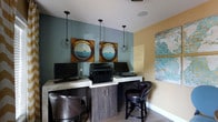 This single-floor business center offers a thoughtfully designed workspace that balances functionality with an inviting aesthetic. The room features a nautical-inspired décor theme, highlighted by a pair of distinctive circular porthole-style art pieces mounted on a cool slate-blue accent wall, evoking a coastal ambiance. A warm golden-yellow wall complements the cooler tones, adding visual warmth and contrast to the space. Overhead, a series of industrial-style cage pendant lights hang from the ceiling, casting a warm, focused glow over the workstations below. The built-in workstation counter runs along the wall and is finished with a light stone-style surface, supported by dark wood cabinetry beneath. Multiple computer workstations are set up along this counter, accompanied by a shared printer and scanner for added convenience. Richly upholstered dark leather swivel chairs provide comfortable seating at each station. Light wood-toned flooring ties the space together, while an abstract blue-toned canvas artwork on the adjacent wall adds a final artistic touch. The overall ambiance is professional yet welcoming, making it a well-equipped and aesthetically refined space for focused work.