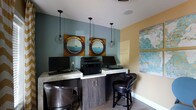 This single-floor business center offers a thoughtfully designed workspace that balances functionality with an inviting aesthetic. The room features a nautical-inspired décor theme, highlighted by a pair of distinctive circular porthole-style art pieces mounted on a cool slate-blue accent wall, evoking a coastal ambiance. A warm golden-yellow wall complements the cooler tones, adding visual warmth and contrast to the space. Overhead, a series of industrial-style cage pendant lights hang from the ceiling, casting a warm, focused glow over the workstations below. The built-in workstation counter runs along the wall and is finished with a light stone-style surface, supported by dark wood cabinetry beneath. Multiple computer workstations are set up along this counter, accompanied by a shared printer and scanner for added convenience. Richly upholstered dark leather swivel chairs provide comfortable seating at each station. Light wood-toned flooring ties the space together, while an abstract blue-toned canvas artwork on the adjacent wall adds a final artistic touch. The overall ambiance is professional yet welcoming, making it a well-equipped and aesthetically refined space for focused work.